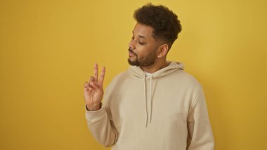 Afro-Amerikan bir adam sarı arka planda hoş bir gülümsemeyle barış işareti yapıyor.