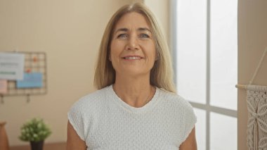 Gülümseyen orta yaşlı bir kadın minimalist dekorla aydınlık bir oturma odasında güvenle duruyor.