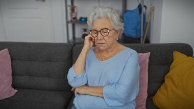 Düşünceli, gözlüklü yaşlı bir kadın rahat bir oturma odasında, bir kanepede dinleniyor, yalnızlığı ve yansımayı somutlaştırıyor..