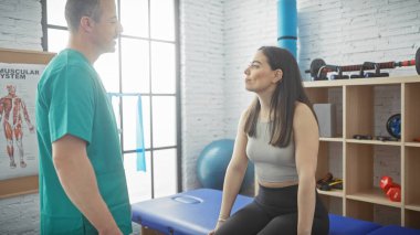 Bir kadın hasta, erkek bir fizyoterapistle egzersiz ekipmanlarıyla dolu, parlak bir rehabilitasyon odasında ilişki kurar..