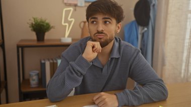 Sakallı düşünceli genç bir adam evde oturup modern bir oturma odasına bakıyor..