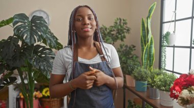 Saçları örgülü gülümseyen Afrikalı bir Amerikalı kadın yeşil bir çiçekçide önlük takıyor..