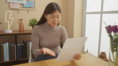 Genç Asyalı bir kadın evde dikkatlice bir belge okuyor, iyi aydınlatılmış bir odada duygularını gösteriyor..
