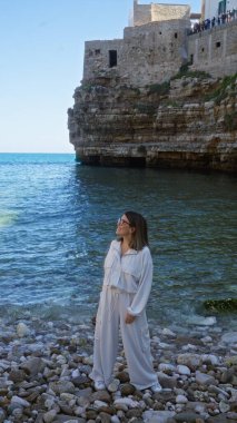 A young, beautiful hispanic woman stands on the beach in polignano a mare, puglia, italy, with the mediterranean sea and historic seaside buildings in the background. clipart