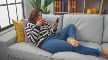 Rahat, orta yaşlı bir kadın, rahat bir oturma odasındaki kanepede telefonuna bakarken kahve içer.