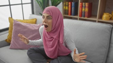 Kapalı bir koltukta oturan tesettürlü şaşkın kadın, arka planda bir kitaplık ve yastıkla şaşkınlığını dile getiriyor..