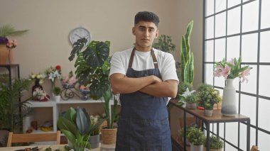 Çapraz kollu kendinden emin bir adam kapalı bir çiçekçide dikiliyor. Etrafı bitkiler ve çiçeklerle çevrili..