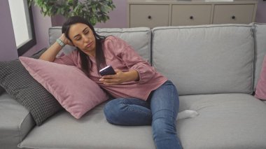 Rahat genç bir kadın evdeki bir kanepede uzanır, akıllı bir telefonla günlük kıyafetlerini giyer..