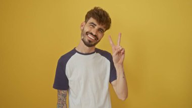 Sakallı, neşeli, genç, İspanyol bir adam canlı sarı bir arka plan üzerinde barış işareti yapıyor..