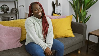 Gülümseyen Afro-Amerikan kadın kırmızı örgülü gri bir kanepede oturuyor rahat bir oturma odasında..