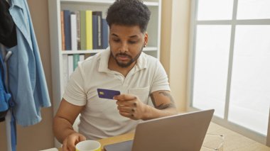 Odaklanmış siyah bir adam, rahat bir oturma odasında dizüstü bilgisayarla çalışırken kredi kartını inceliyor..