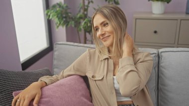 Güzel, genç, sarışın bir kadın rahat bir oturma odasında oturuyor, gülümsüyor ve yumuşak yastıklarla modern bir koltukta dinleniyor..