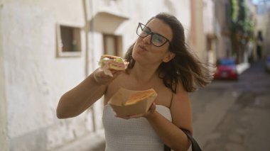 Genç bir İspanyol kadın güneşli bir günde Gelibolu, Puglia, İtalya 'nın manzaralı sokaklarında yürürken buccia sandviçi yer..