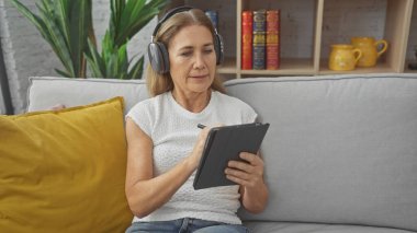 Normal kıyafetli orta yaşlı bir kadın modern oturma odasındaki bir kanepede tablet kullanırken kulaklıkla müzik dinliyor..