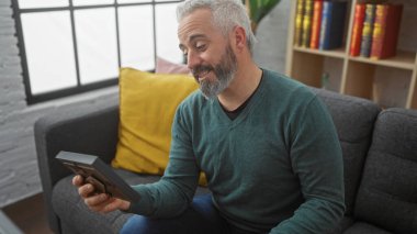 Olgun sakallı bir adam, elinde tablet, kitaplık ve modern dekorla evde otururken gülümsüyor..