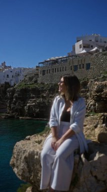 Genç ve güzel bir İspanyol kadın polignano 'da sahile ve denize bakan kayalık bir uçurumda oturuyor. Bir kısrak, puglia, İtalya. Arkasında parlak mavi bir gökyüzünün altında tarihi binalar var..