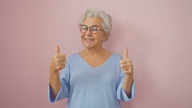 A cheerful elderly woman with grey hair gives thumbs up in front of a pink background, expressing positivity and happiness. clipart