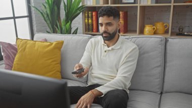 Modern bir oturma odasında elinde uzaktan kumandayla televizyon izleyen sakallı genç bir adam.