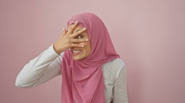 Pembe tesettürlü genç bir kadın izole pembe bir arka planda, parmaklarının arasından oyuncu bir şekilde gözetliyor.