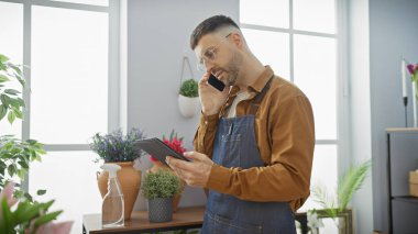 Yakışıklı bir İspanyol, çiçekçi dükkanında telefon ve tabletle birden fazla iş yapıyor. Etrafı canlı bitkilerle çevrili..