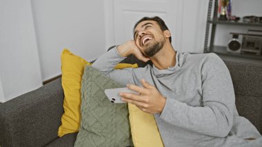 Oturma odasındaki kanepede akıllı telefon kullanan sakallı, gülen genç İspanyol adam.