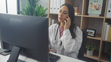 Klinikteki profesyonel bir İspanyol kadın doktor telefonda konuşurken bilgisayar ekranına odaklanır..