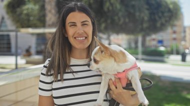 Şehir parkında Chihuahua köpeğini tutan genç bir İspanyol kadın bu açık şehir sokak sahnesinde içten bir gülümseme ile gülümsüyor..