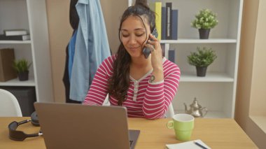 Genç İspanyol kadın telefonda konuşuyor, ahşap bir masada dizüstü bilgisayarla otururken sinsice gülümsüyor, iyi organize edilmiş bir odada..