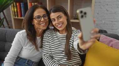 İki kadın, muhtemelen anne ve kız, keyifli bir selfie anı paylaşıyorlar. Oturma odasındaki kanepede..