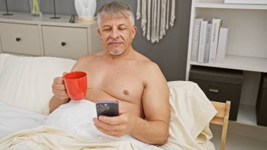 Olgun bir adam, çağdaş bir yatak odasında akıllı telefonunu karıştırırken sabah kahvesini yatakta içmekten hoşlanır..