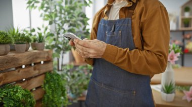 Önlüklü İspanyol bir adam kapalı bir çiçekçide canlı bitkilerin arasında akıllı telefon kullanıyor..