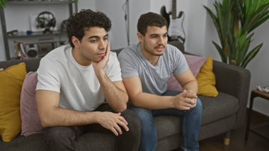 Modern bir oturma odasındaki kanepede iki genç adam düşünceli bir şekilde oturuyor..