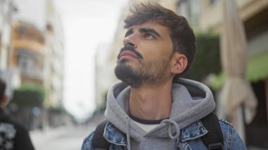 Sakallı genç bir Hispanik adam, açık bir şehir caddesinde dikilirken derin derin bakıyor..