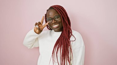 Pembe arka planda örgü ve gözlüklü gülümseyen Afrikalı Amerikalı kadın barış işareti gösteriyor