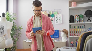 Sakallı genç bir adam mankenlerle ve dikiş malzemeleriyle dolu renkli bir terzi dükkanındaki tablete odaklanmış..