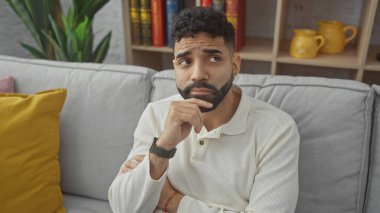 Sakallı, düşünceli, genç, İspanyol bir adam rahat bir dairede oturuyor..