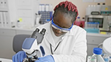 Örgü takan Afro-Amerikalı bir kadın laboratuarda mikroskop kullanarak bir örneği analiz ediyor..