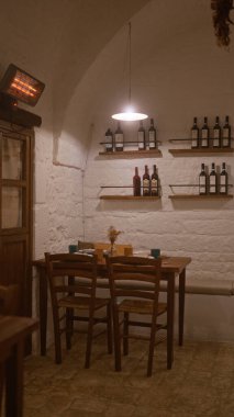 Kapalı bir İtalyan restoranı, rahat bir masa düzeneği, kırsal tuğla duvarlar, ahşap sandalyeler, raflar dolusu şarap şişeleri ve loş ışık, sıcak bir ortam yaratıyor..
