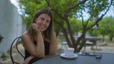 Siyah bluzlu genç İspanyol kadın İtalya 'da bir kafede oturuyor, elinde bir fincan kahveyle kameraya gülümsüyor..