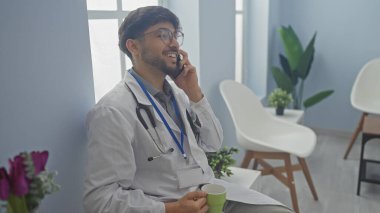 Doktor üniformalı gülümseyen adam klinikteki bekleme odasında kahve fincanı ve arka planda bitkilerle akıllı telefon kullanıyor.
