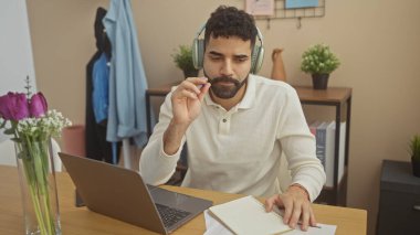 Ev ofisinde çalışan, kulaklık takan ve elinde kalem tutan odaklı, sakallı bir İspanyol..