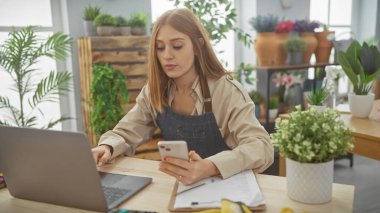 Odaklanmış genç bir kadın, bilgisayarlı ve akıllı telefonlu çoklu görevlerle dolu, iyi aydınlatılmış bir ev ofisinde çalışıyor.