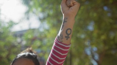 A young woman raises her tattooed fist in solidarity, outdoors surrounded by green foliage. clipart