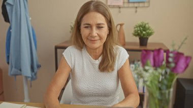 Olgun, beyaz bir kadın. Kapalı alanlarda, bitkilerle, modern dekorla ve günlük tarzıyla gülümsüyor..