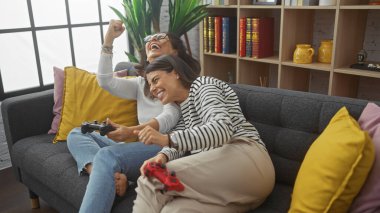 İki kadın, rahat bir oturma odasında kanepede gülüp oyun oynuyorlar..