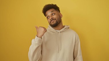 Yakışıklı siyahi adam sarı bir arka planın önünde başparmağını yana doğru gösteriyor. Kapüşonlu ve düşünceli bir ifade takınıyor..