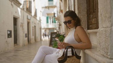 Güneş gözlüklü genç, güzel, İspanyol bir kadın locorotondo, puglia, İtalya 'nın eski kasabasının sokaklarında güneşli bir günde tarihi beyaz binaların etrafında akıllı telefon kullanıyor..