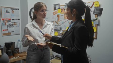 İki kadın müfettiş, bir polis karakolunun suç analiz odasındaki delilleri analiz ediyor, fotoğraflar ve dava dosyalarıyla çevrili..