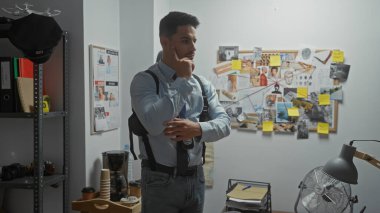 Sakallı, düşünceli, genç, İspanyol bir adam dedektifin ofisinde duruyor ve bir kanıt panosunu inceliyor..