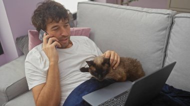 Genç bir adam telefonda kedi beslerken ve rahat bir oturma odasında dizüstü bilgisayar kullanırken konuşuyor..